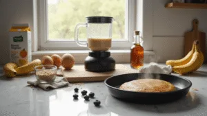 A bright morning kitchen scene featuring a marble countertop with a professional blender filled with protein pancake batter, surrounded by fresh ingredients like whole eggs, a ripe banana, almond milk, measuring cups, coconut flour, vanilla protein powder, and golden flaxseeds. A golden-brown pancake in a cast iron pan emits steam, alongside scattered blueberries and a bottle of maple syrup, all illuminated by soft natural sunlight. The atmosphere is warm and inviting, embodying a modern minimalist aesthetic.