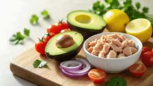 Cinematic close-up of a rustic wooden cutting board with halved avocado, flaked tuna in a white bowl, scattered cherry tomatoes, red onion rings, halved lemon, and fresh herbs, all in soft natural daylight.