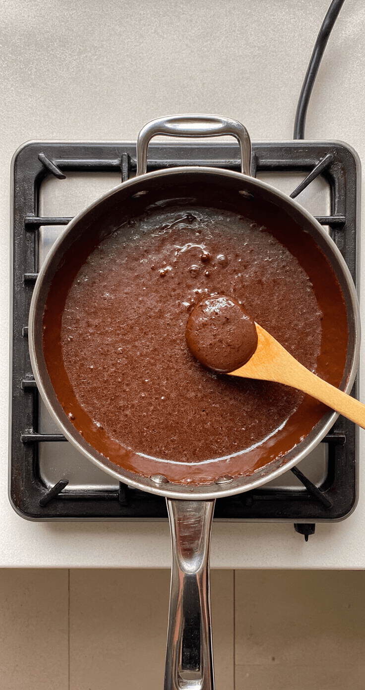 Keto Kokos Fudge – Suikervrij, Romig en Klaar in 30 Minuten Close-up van een antiaanbak steelpan op laag vuur met een glanzend chocolade-kokosmengsel dat pruttelt, met een houten lepel ernaast en luchtige stoom in warm keukenlicht.