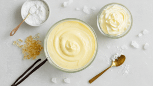 Keto Vanillepudding: Romig, Suikervrij en Klaar in 10 Minuten Overhead shot of a pristine white marble kitchen countertop with glass bowls of erythritol crystals, golden vanilla extract, fresh whipped cream in a crystal jug, and powdered gelatin in a porcelain ramekin, accompanied by a copper whisk, soft natural lighting, subtly blurred modern Dutch kitchen in the background, warm inviting atmosphere, rich textures, cinematic depth.