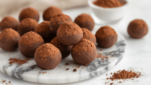 Keto Romige Chocolade Truffels – Suikervrij en Simpel Close-up van handgemaakte chocolade truffels bestrooid met cacaopoeder op een marmeren werkblad, met gesmolten chocolade en ingrediënten in de achtergrond, omgeven door warme verlichting.