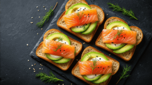 Gerookte Zalm met Avocado en Citroenmayonaise Bovenaanzicht van vier geroosterde broodsneetjes belegd met citroenmayonaise, avocado en gerookte zalm, op een zwart leisteen serveerblad, omringd door verse dille, zwarte sesamzaadjes, en een gehalveerde citroen met vintage lepel.