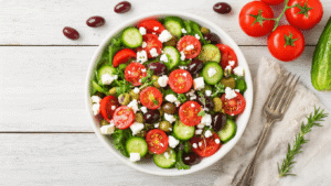 Griekse Salade met Feta – Vers, Makkelijk en Klaar in 15 Minuten Cinematische overheadfoto van een authentieke Griekse salade met feta, tomaten, komkommer en olijven, geserveerd in een witte keramische schaal op een rustieke houten tafel, vergezeld van een glazen kom met dressing en warme natuurlijke belichting.
