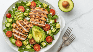 Kip-Avocado Salade: Romig, Fris en Klaar in 20 Minuten Dramatisch zijaanzicht van een rustieke schaal met kleurrijke kip-avocado salade, gegrilde kipfilet, romaine sla, cherrytomaat, komkommer, feta en een gouden limoen-mosterd dressing, verlicht door zachte natuurlijke verlichting op marmeren ondergrond.
