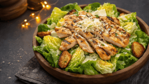 Keto Caesar Salade (Zonder Croutons) Keto Caesar salade met romaine sla, gegrilde kip, Parmezaanse kaas en Caesar dressing, geserveerd in een rustieke houten kom op donker leisteen, met een halve citroen en versgemalen zwarte peper.
