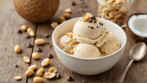 Keto Ijs van Kokosmelk en Pindakaas: Romig, Suikervrij en Verrassend Makkelijk Cinematic overhead view of a rustic wooden table featuring a white ceramic bowl filled with creamy keto coconut peanut butter ice cream, topped with chopped roasted peanuts and sugar-free chocolate chips, alongside an open can of full-fat coconut milk and a glass jar of 100% peanut butter, bathed in soft warm light.